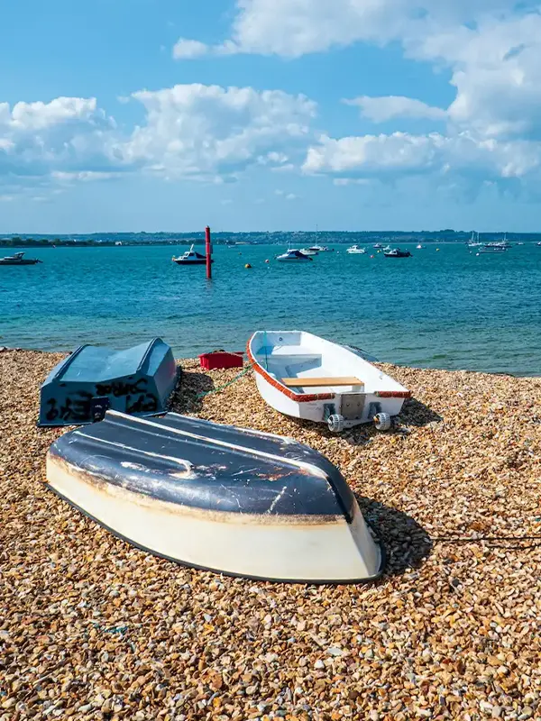 Kleine Boote auf einem Kiesstrand mit Segelbooten vor der Küste unter blauem Himmel.