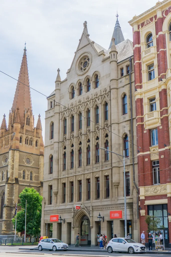 Ein Gebäude der Torrens University an der Flinders Street in Melbourne. Die gotische Sandsteinfassade zieht sich über mehrere Stockwerke nach oben, am Eingang fallen zwei knallrote Schilder der Universität sofort ins Auge. Links im Bild ragen die Türme der St. Paul's Cathedral hinein.