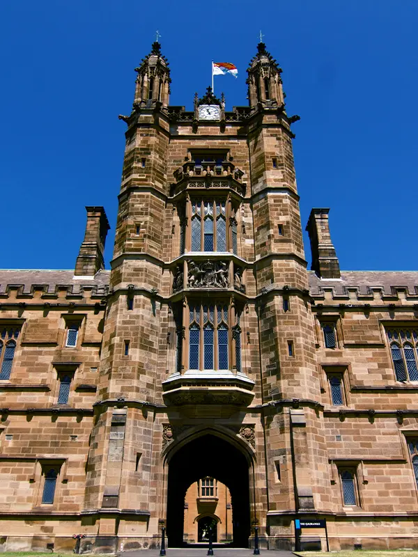 Nahaufnahme des gotischen Sandstein-Torturms am Quadrangle der University of Sydney mit Zwillingstürmen, einer Uhr und der Universitätsflagge über dem Bogendurchgang; ein führendes Ziel für internationale Studierende in Australien.