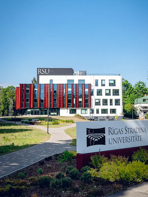 Ein modernes, weißes RSU-Gebäude mit roten vertikalen Paneelen und dem Schriftzug „Rīgas Stradiņa Universitāte“, umgeben von grünen Gärten an einem sonnigen Tag