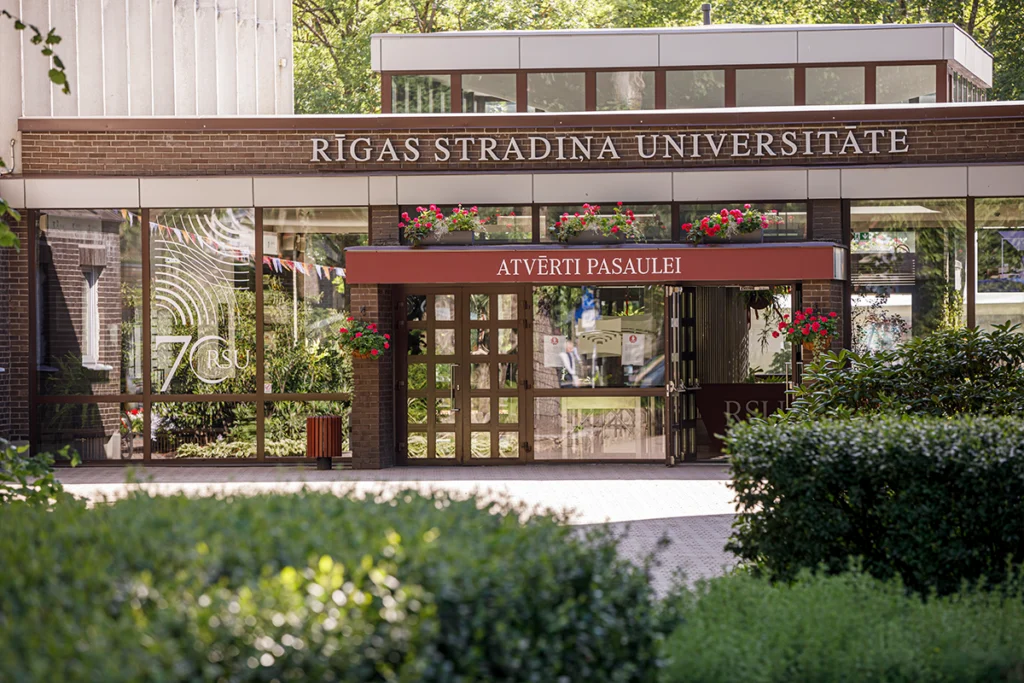 Haupteingang der Stradins-Universität in Riga mit dunkler Backsteinfassade, RSU-Schild, roten Geranien und dem Motto „Atvērti Pasaulei“ (offen für die Welt)