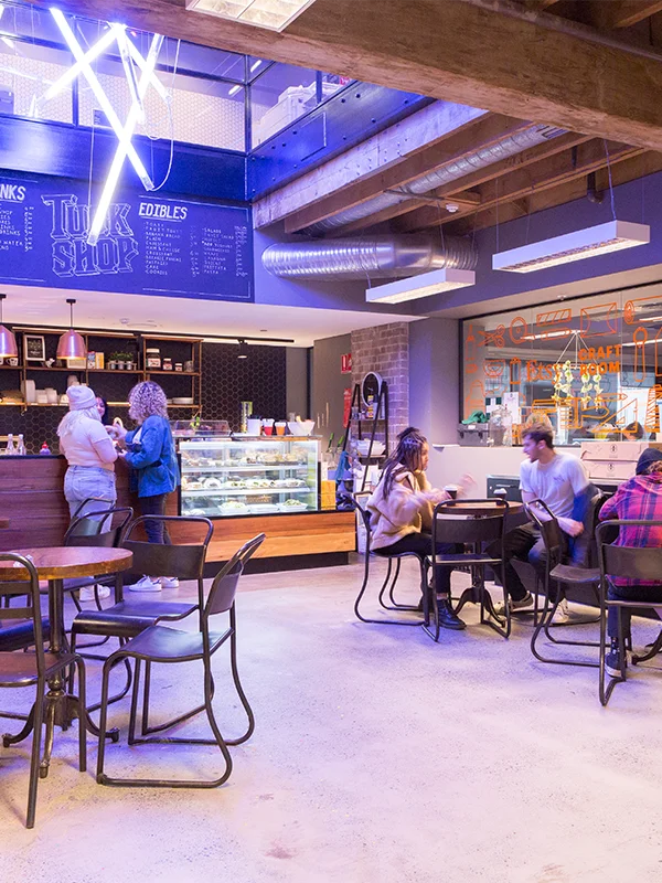 Studierende an Tischen im Industriestil im Tuck Shop Café der Torrens University Adelaide mit blauem Neonlicht und freiliegenden Holzbalken an der Decke