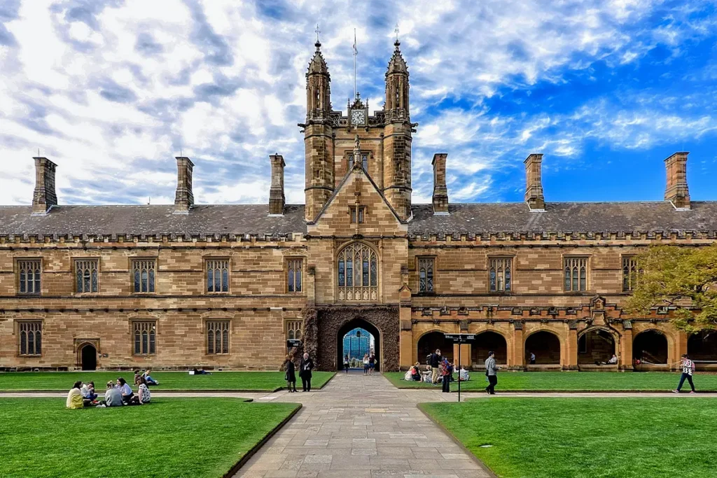 Weitwinkelansicht des Quadrangle-Innenhofs der University of Sydney mit Studierenden auf dem Rasen, dem gotischen Sandstein-Uhrturm im Hintergrund und Kolonnaden rechts; repräsentiert das Campusleben an einer der besten Universitäten Australiens für internationale Studierende.