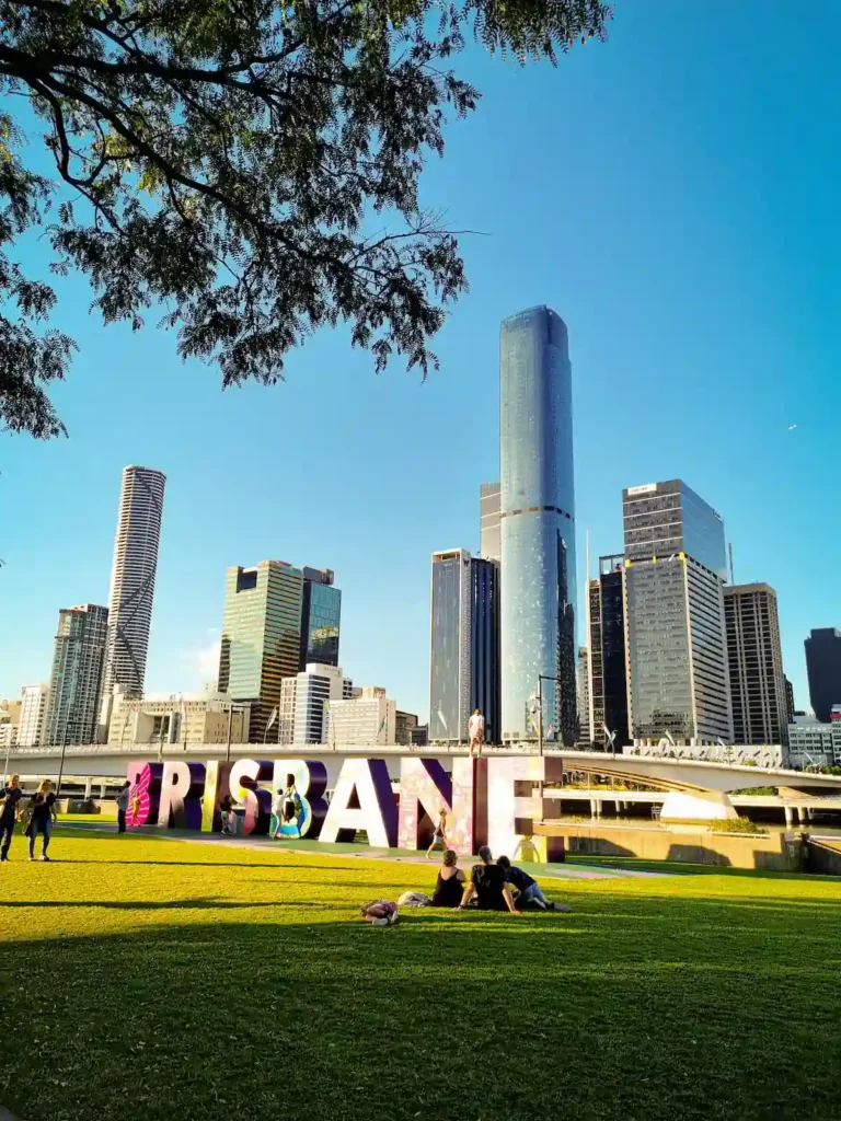 Bunter Brisbane-Schriftzug im Vordergrund mit der Stadtsilhouette und blauem Himmel im Hintergrund